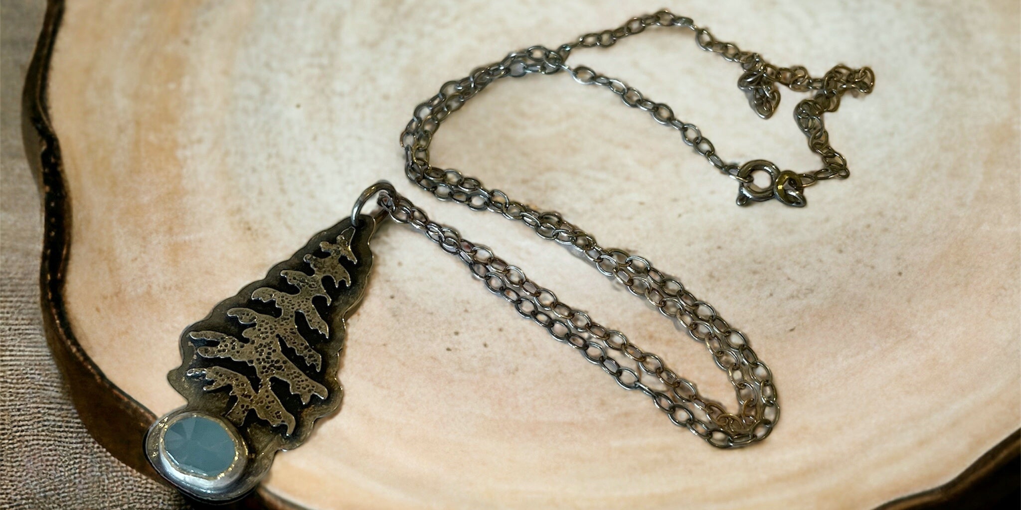 a necklace with a silver evergreen tree  and an aquamarine rests on a plate on a light tan background