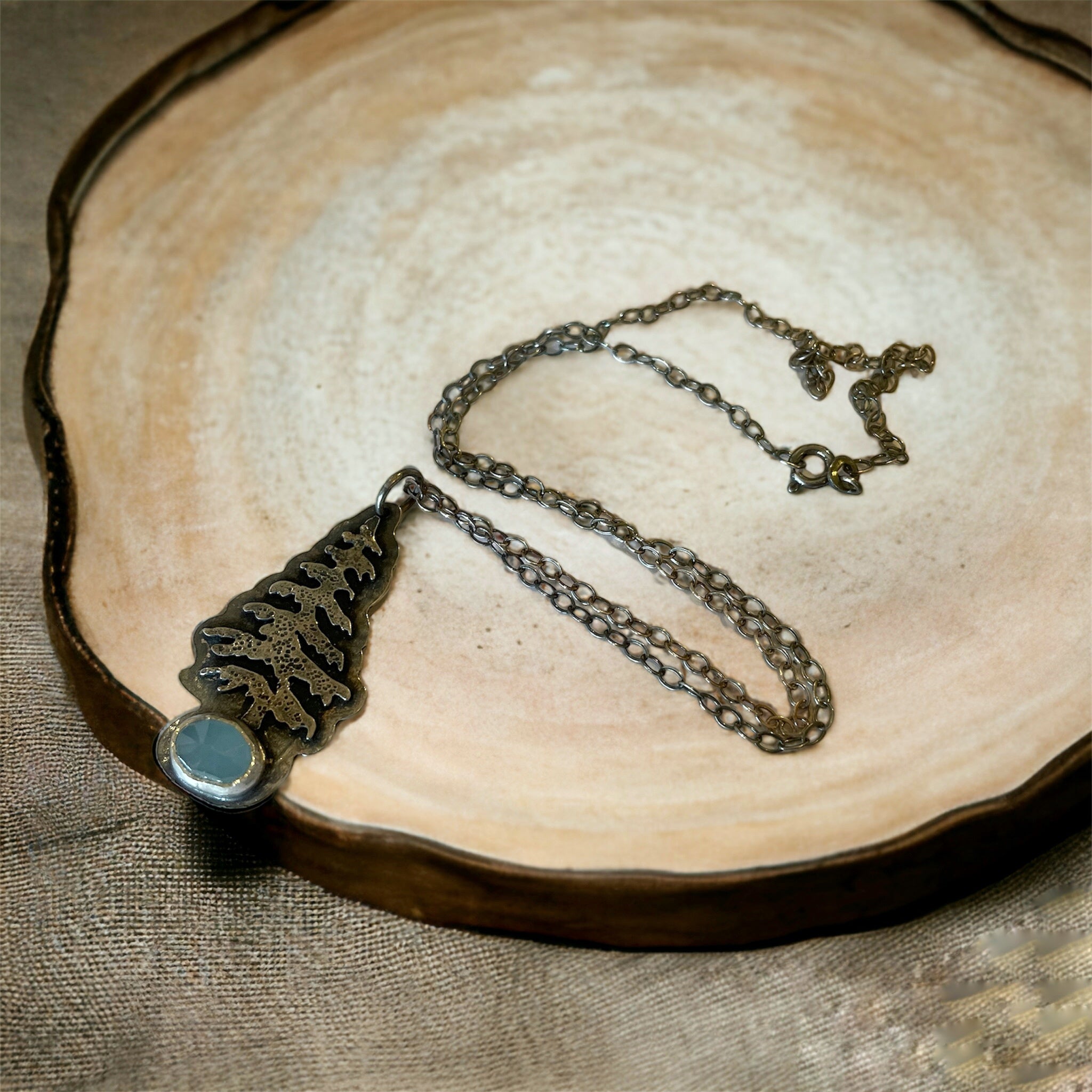 a necklace with a silver evergreen tree  and an aquamarine rests on a plate on a light tan background